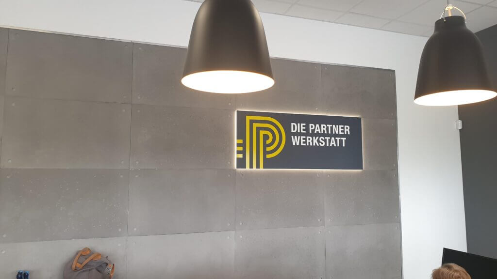 Innenbeschilderung „Die Partnerwerkstatt“ mit gelbem Logo an einer grauen Wand